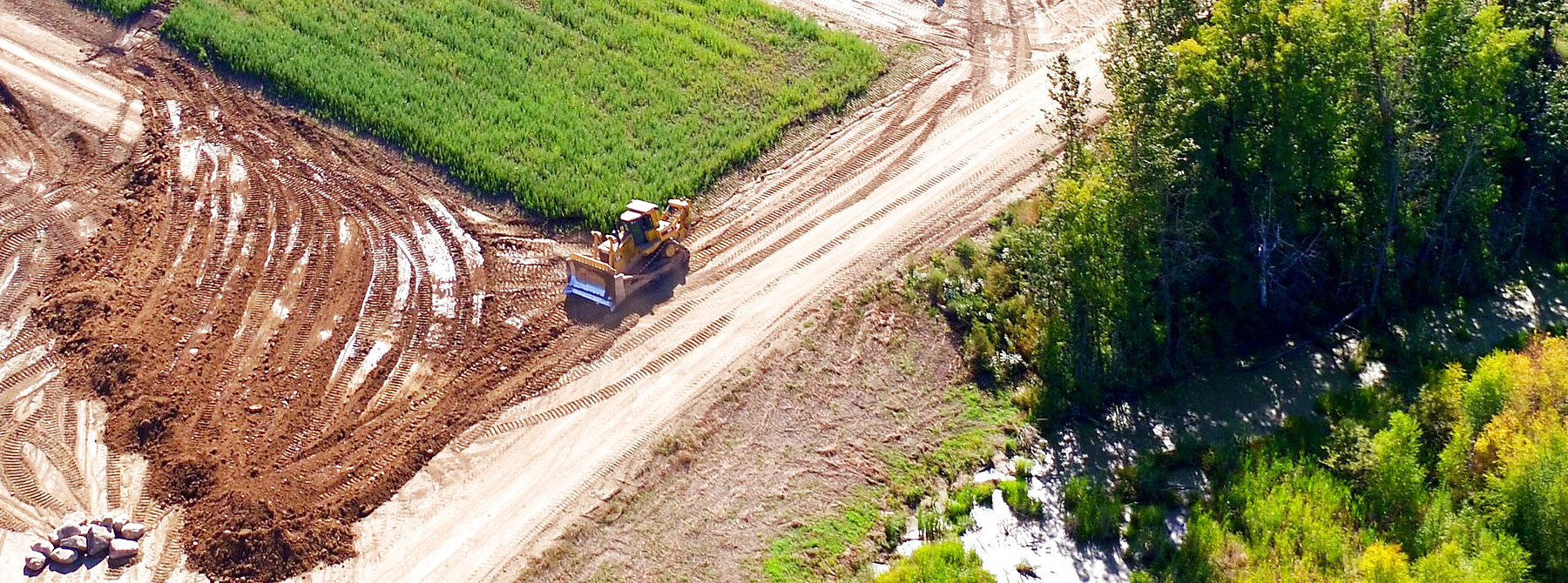 Worked ground from the air: a machine cutting a corridor between a treeline and a field, windrowed spoil along one side and machine passes across the cut floor.