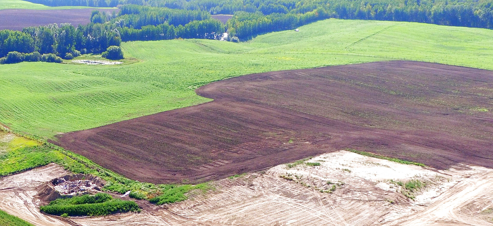 Fields on ranch land seen from the air: a green field and a ploughed one meeting at a tree line, and the edge of a stripped pad below them.
