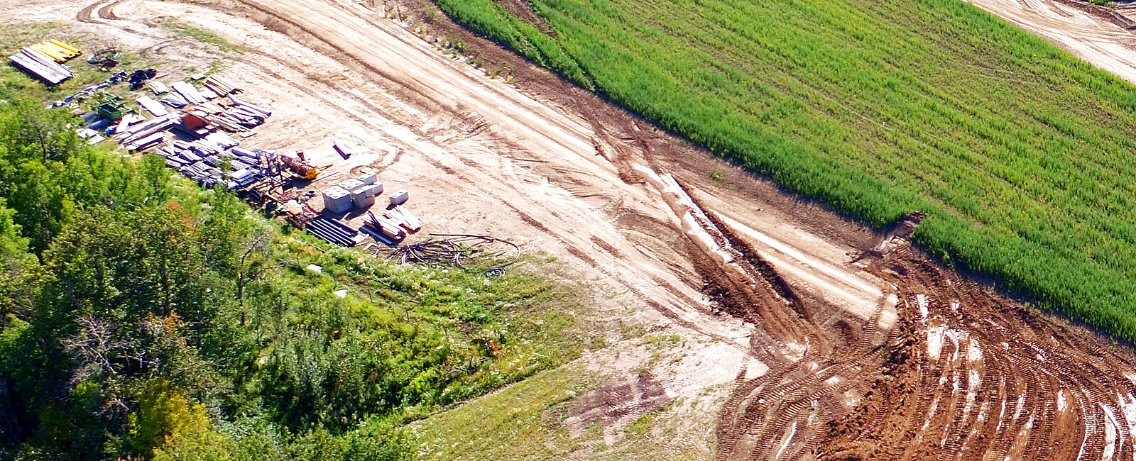 A laydown yard at the edge of earthworks, seen from the air: stacked pipe, timber and concrete block on the grass beside the haul road, the cut beyond it.