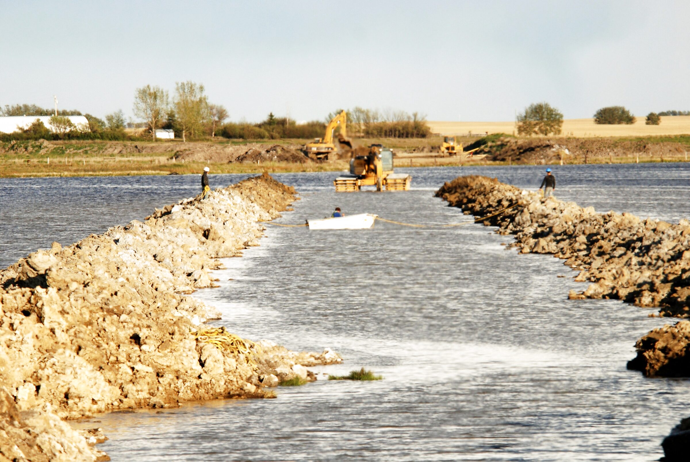 A rock berm running out through standing water, built up from the bed, with worked ground on the bank behind it.